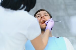 A dentist wearing gloves examines a male patient’s mouth using dental tools during a checkup.