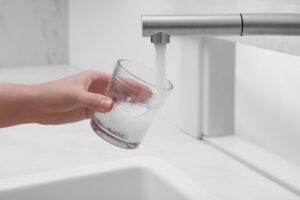 A hand holds a clear glass under a modern kitchen faucet as water pours into the glass, with a white sink below.
