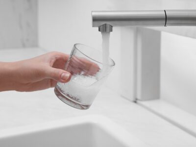 A hand holds a clear glass under a modern kitchen faucet as water pours into the glass, with a white sink below.
